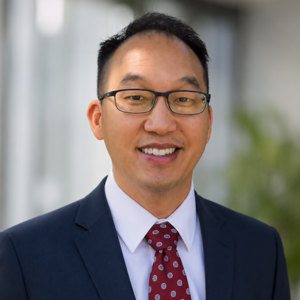 Smiling man in a navy suit and red patterned tie, wearing glasses, outdoor background.